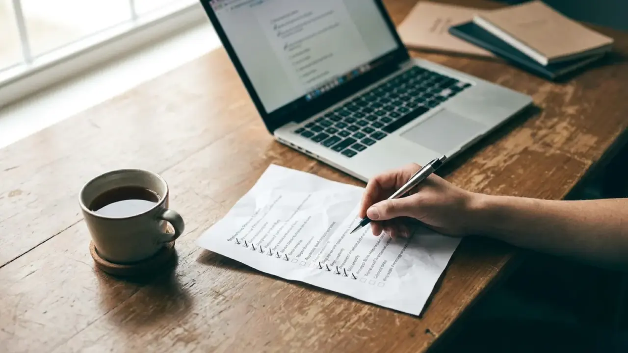 A laptop, a ticked printed checklist and a cup of tea on a desk, seen from above