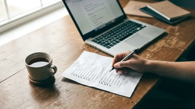 A laptop, a ticked printed checklist and a cup of tea on a desk, seen from above